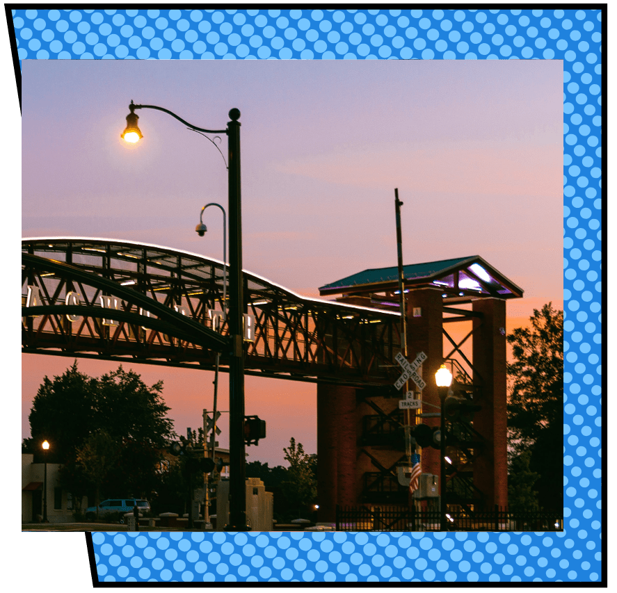 A metal bridge arches over a street in Acworth, with warm streetlights illuminating the scene. The sky is a gradient of pink and orange, creating a serene atmosphere.