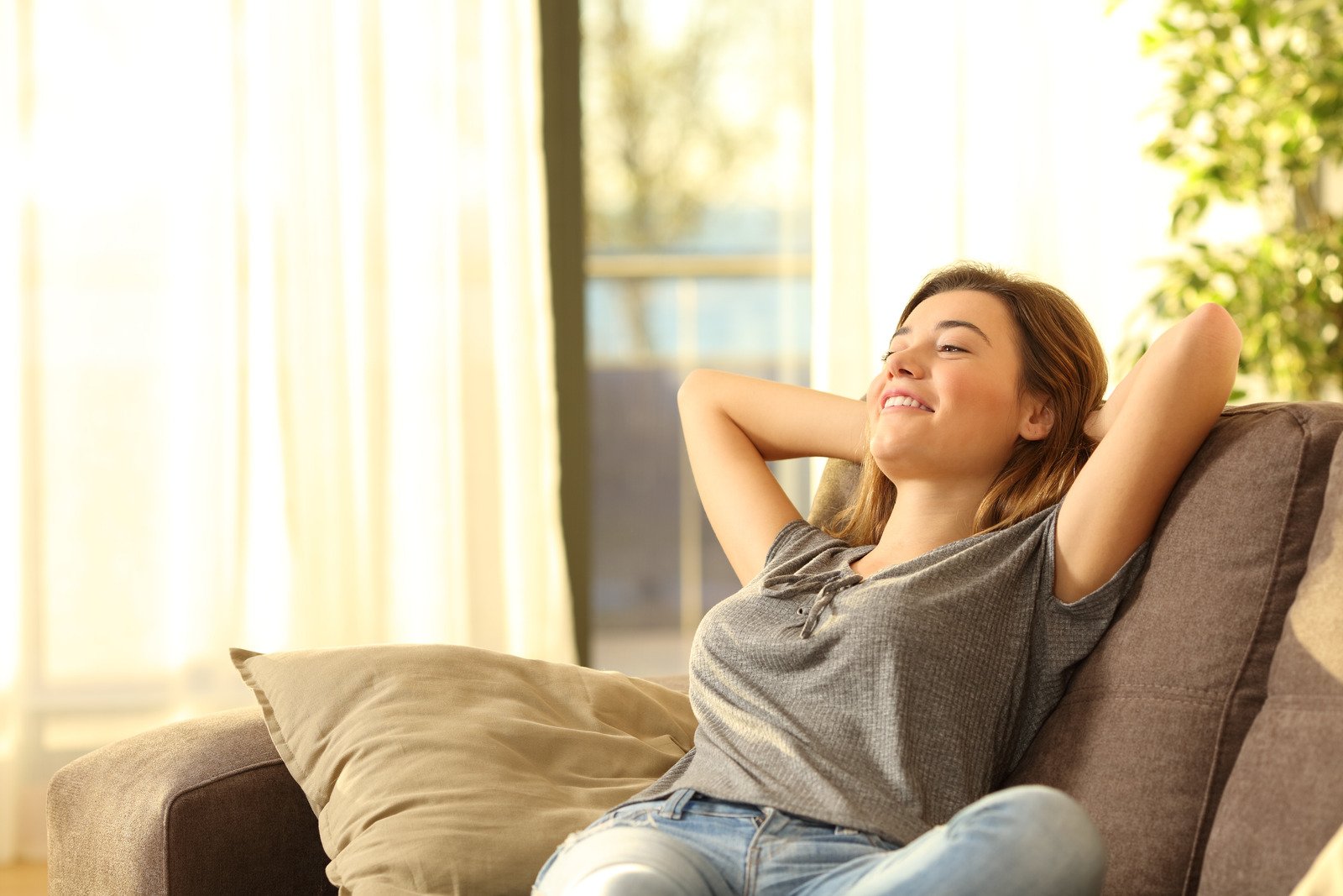 a young women leaning back into a sofa with the sun shining on her, her eyes closed and a smile on her face