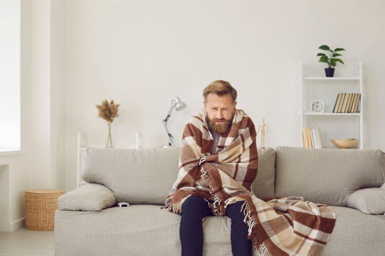 man wrapped in a throw blanket sitting on his couch in the living room freezing because of a broken furnace