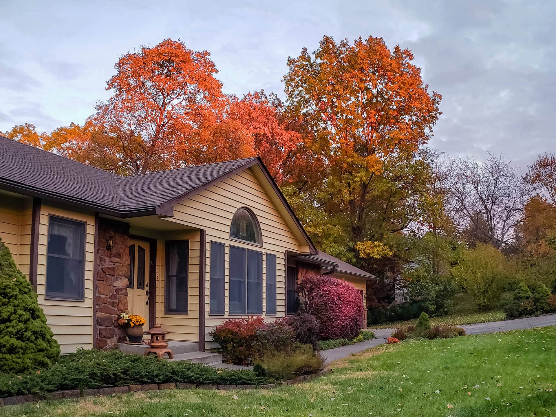 A cozy house with light yellow siding and large windows is surrounded by lush autumn foliage