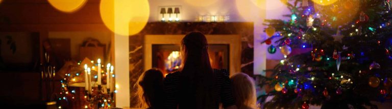 A family gazes at a Christmas tree adorned with colorful lights and ornaments. A cozy fireplace glows gently, creating a warm, festive atmosphere.