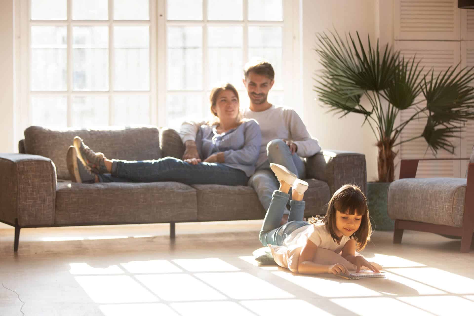 A family relaxing in a bright, sunlit living room with a young girl drawing on the floor, illustrating a comfortable home environment and the ideal time when to turn on AC to maintain indoor air quality and temperature.