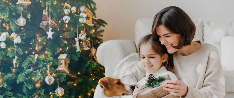 A mother and daughter share a joyful moment with their dog by a decorated Christmas tree. The scene is cozy and festive, evoking warmth and happiness.