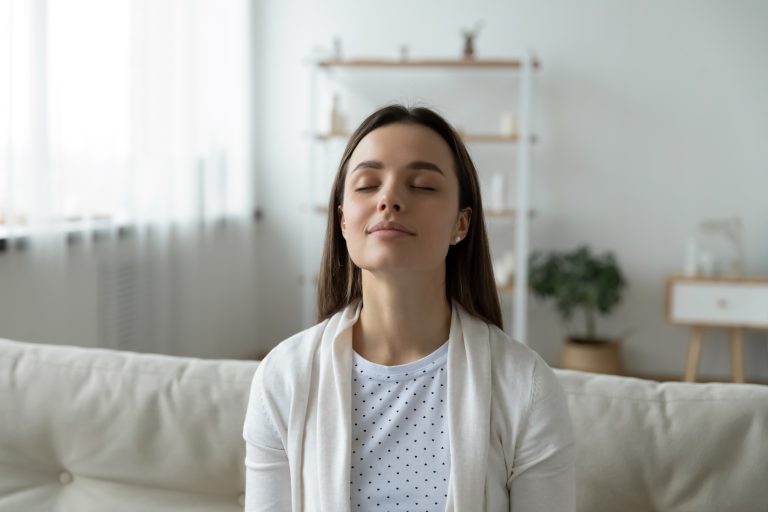 A woman sits on a sofa with her eyes closed, appearing calm and relaxed. The room is softly lit, featuring blurred shelves and a plant in the background.