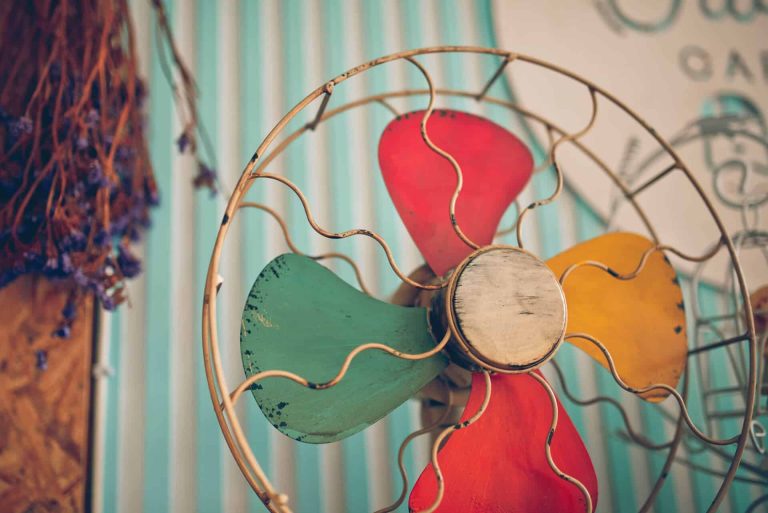 Vintage fan with colorful blades—red, green, yellow—encased in a wire frame. Background features striped wallpaper and dried flowers, evoking nostalgia.