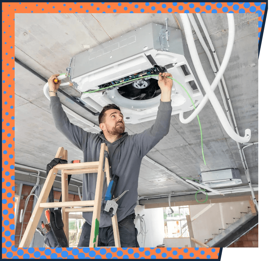 A man stands on a wooden ladder fixing a ceiling air conditioning unit, focusing intently on wiring. The setting is a partially constructed room.
