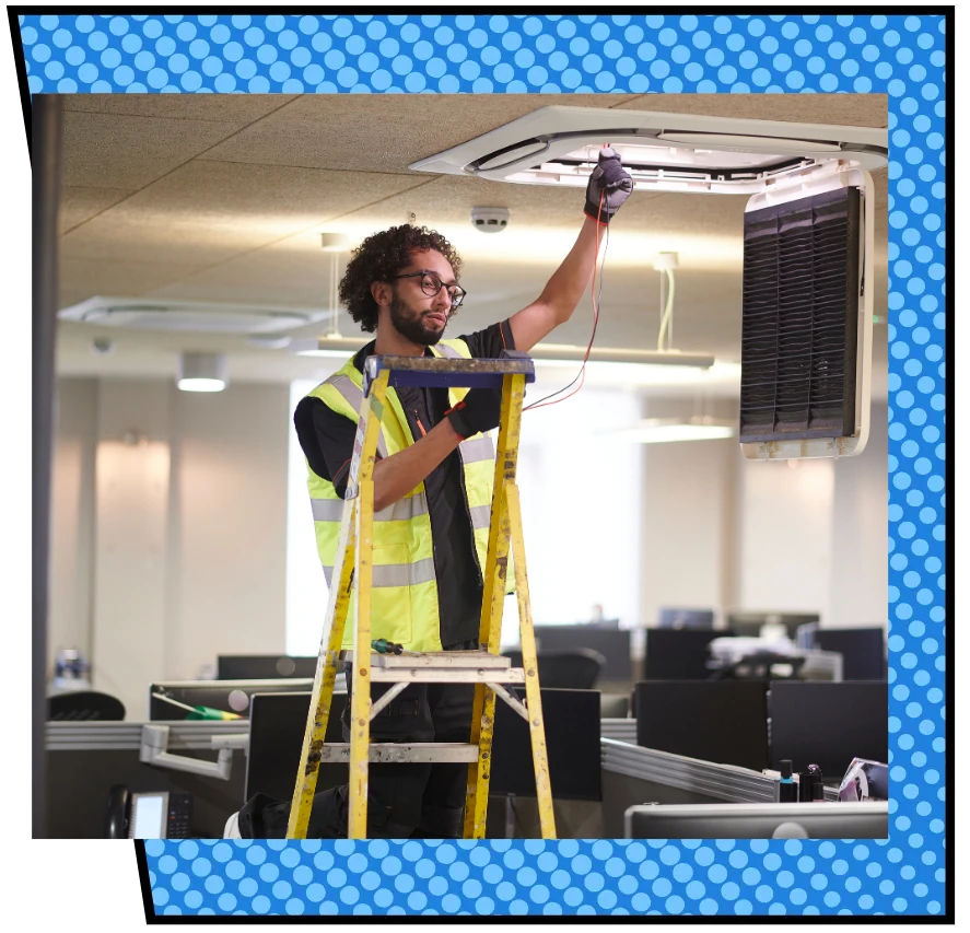 A maintenance worker in a safety vest and gloves stands on a ladder adjusting a ceiling air vent in a brightly lit, modern office space.