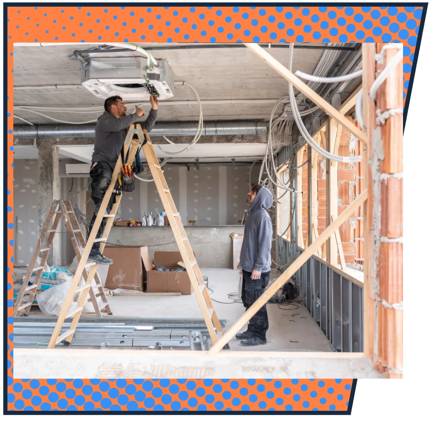 Two workers install a ceiling unit in an under-construction room. One stands on a ladder, the other observes. The atmosphere is focused and industrious.