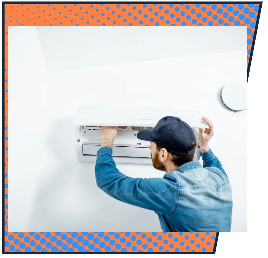 A man in a denim jacket and cap repairs a wall-mounted air conditioner. The room is bright, conveying a clean, focused work atmosphere.