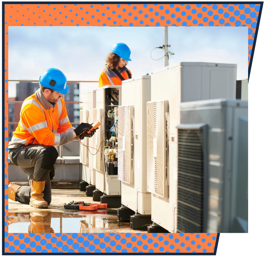 Two construction workers in orange vests and blue helmets servicing rooftop HVAC units. Bright, sunny day with urban skyline background.