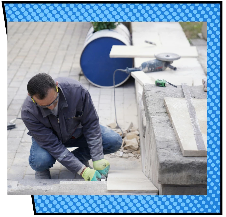 A man in work gloves and goggles kneels on a paved surface, cutting stone. Nearby are tools and a blue barrel, set against a polka dot border.
