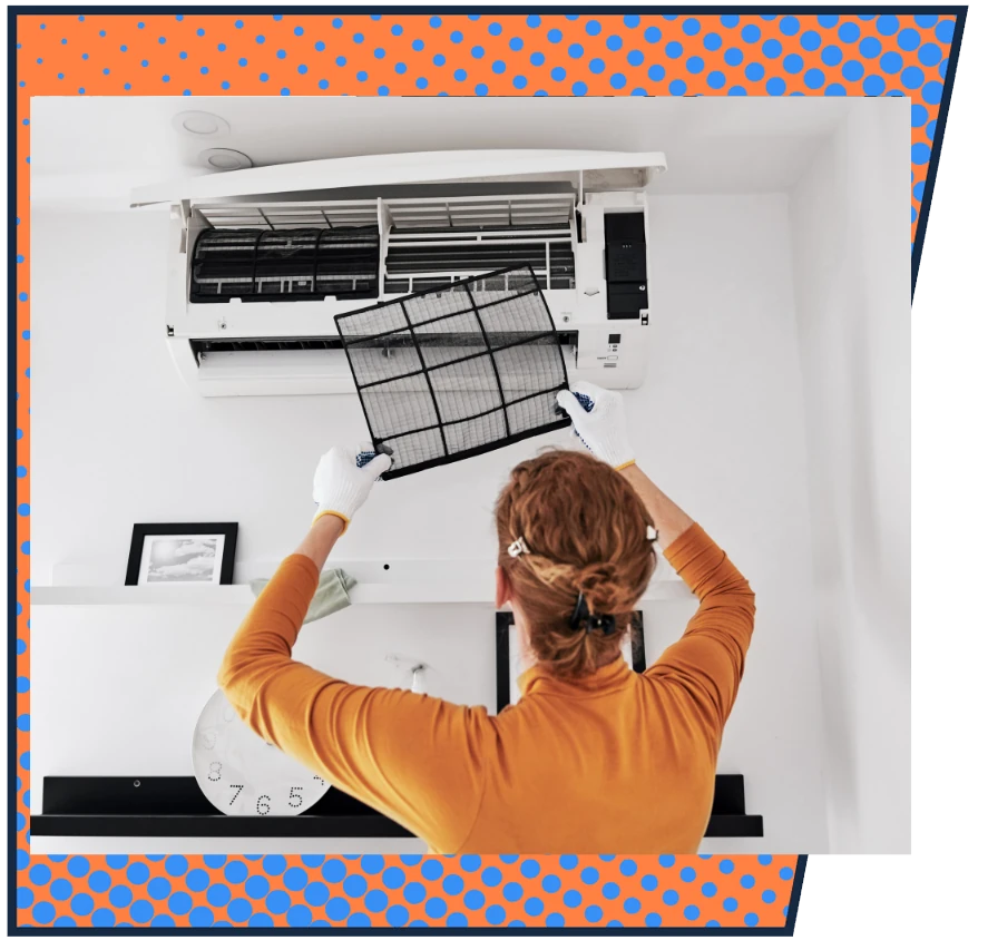 A person in an orange sweater and gloves cleans an air conditioner filter in a modern room. The action conveys cleanliness and maintenance.