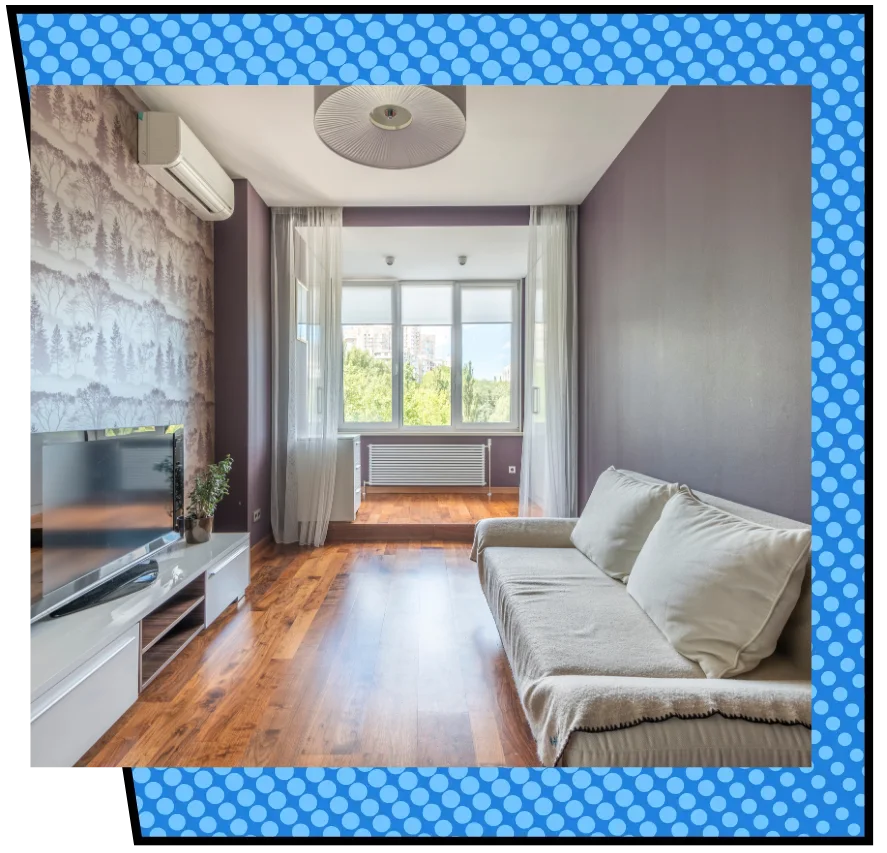 Stylish living room with wood floors, a sofa, and a TV. Large windows offer natural light, with sheer curtains and a view of trees outside.